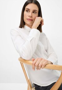 Weißes, langärmliges Hemd mit Knopfleiste, weicher Textur, minimalistischem Design und schmalem Kragen, getragen von einer sitzenden Person mit heller Hautfarbe.