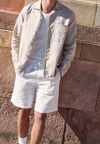 Chemise en lin beige clair avec une poche poitrine unique, portée sur un t-shirt blanc et associée à un short en coton blanc, accessoirisée avec des chaussettes blanches.