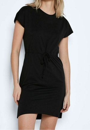 Nainen, jolla on lyhyt musta mekko, jossa on lyhyet hihat ja solmittu vyötärö, seisoo valkoista taustaa vasten.