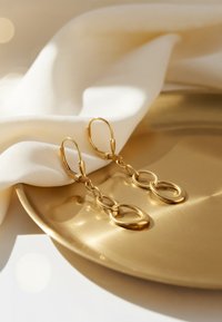 Goldene Creolen mit verbundenen ovalen Formen. Glatte Oberfläche und glänzende Textur vor einem weichen weißen Stoff und goldenem Plattenhintergrund.