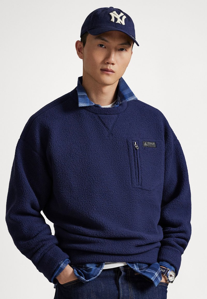 Giovane con indosso un pullover in pile blu navy, camicia a quadri blu, jeans scuri, un berretto da baseball blu navy e un orologio da polso, con le mani nelle tasche.