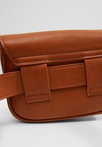 Ruskea nahkavyölaukku, jossa on kirjontayksityiskohtia ja leveä hihna, joka on pujotettu taustan lenkkeihin, levätä valkoisella pinnalla.