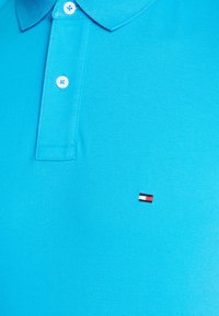 Jasnoniebieska koszulka polo z przewiewnego materiału, z dwoma guzikami oraz małym haftowanym logo w czerwonych, białych i czarnych akcentach.