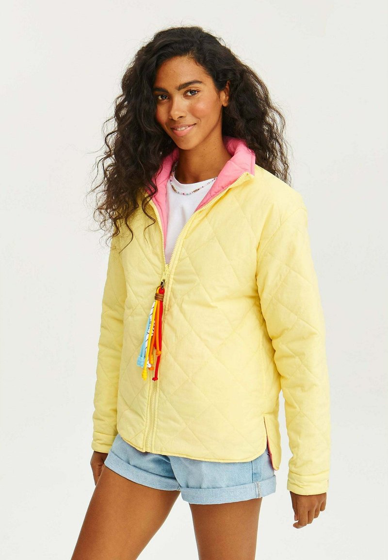 Žlutá prošívaná bunda s růžovým límcem, zipovým uzávěrem a vícebarevnými třásněmi. Nosená přes bílé tričko a džínové šortky, které mají boční rozparky.