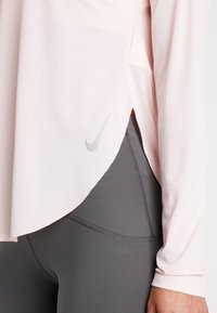 Lichtroze sportieve longsleeve met een gebogen zoom en gespleten zijkanten, gecombineerd met donkergrijze slim-fit leggings met zichtbare naden.