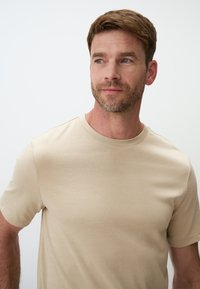 T-shirt beige a girocollo realizzato in morbido cotone, con maniche corte e un design aderente, caratterizzato da una trama liscia e cuciture minime.