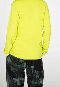 Camisa de manga larga de color amarillo neón con una textura suave; combinada con pantalones negros con un patrón gráfico y detalles de bolsillos laterales.