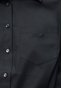 Svart skjorte med knapper og en brystlomme brodert med navnet "Karl" i matchende svart tråd.