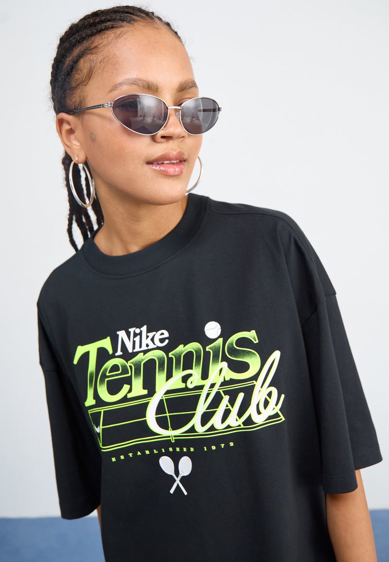 Czarny, oversize t-shirt z grafiką "Nike Tennis Club" w neonowej zieleni i bieli. Model nosi owalne okulary przeciwsłoneczne i duże kolczyki-koła.