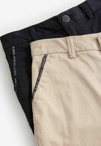 Deux paires de pantalons, l'une noire et l'autre beige, fabriquées en tissu lisse avec des poches latérales et une étiquette de logo visible sur les bords des poches.