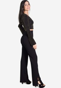 Pantalons noirs évasés avec des fentes à l'ourlet, associés à un haut noir pailleté à manches longues cropped. Le mannequin porte des sandales à talons hauts noires.