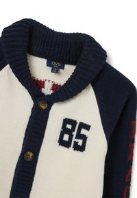 Cárdigan de punto en crema, azul marino y rojo, con cuello de chal, cierre de botones y el número "85" en el frente. Textura suave y mangas en contraste.