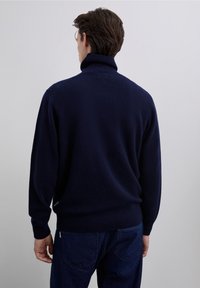 Pull à col roulé en maille bleu marine avec col et poignets côtelés, présentant un tissu texturé et une coupe ample. Jean foncé visible en bas.