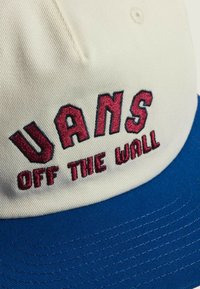 Cappellino con la parte frontale color crema e visiera blu. "UANS OFF THE WALL" è ricamato in rosso sul fronte. Il materiale sembra resistente con cuciture visibili.