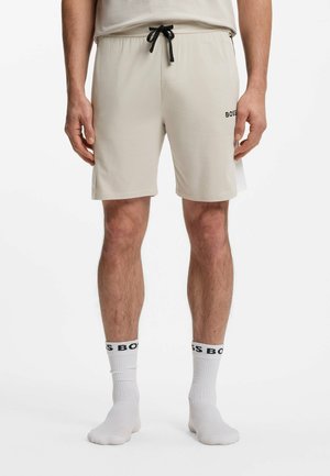 Homme portant un short beige mi-long avec un cordon noir et des chaussettes blanches avec le logo noir "BOSS", debout sur fond blanc.