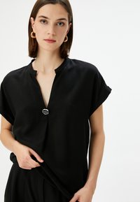 Top nero a maniche corte con scollo a V, caratterizzato da un dettaglio con bottone argentato e una texture liscia. Il design è ampio e minimalista.