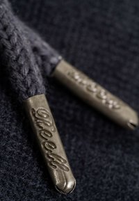 Gray knit shoelaces with metallic aglets featuring embossed lettering. Close-up shows texture and detail of the lace and aglet design.