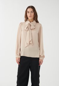 Lys beige bluse med en båndkrave, sammensnørede ærmer og en glat tekstur, parret med sorte bukser.