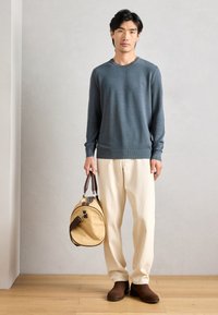 Blå strikket genser, kremfargede bukser, brune sko. Bærer en rund lys brunt veske med mørke detaljer. Modellen står mot en nøytral bakgrunn.