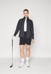 Čierna ľahká bunda s vysokým golierom, biele skrátené tričko, čierne šortky a biele športové topánky; drží golfovú palicu.