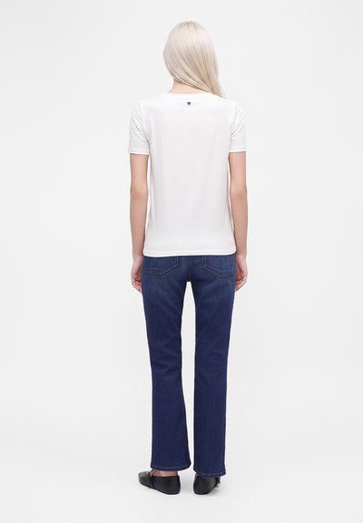 Femme blonde vue de dos portant une chemise blanche à manches courtes, un jean bleu foncé et des chaussures plates noires sur fond blanc.