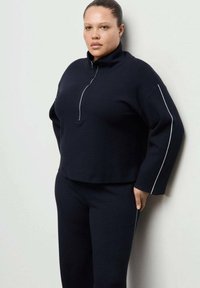 Temno modra majica z visokim ovratnikom in polovičnim zadrgom ter belim poudarkom ob straneh. Kombinirana z ujemajočimi se temno modrimi hlačami, gladke pletene teksture.