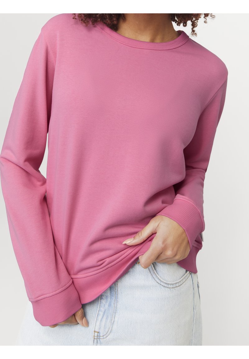 Femme aux cheveux bouclés portant un sweat-shirt rose à manches longues et un jean bleu clair, tenant l'ourlet du sweat-shirt d'une main.