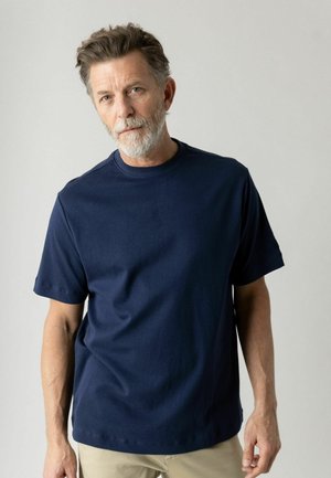 Muž stredného veku so sivou bradou a vlasmi, oblečený v tmavomodrom tričku s krátkym rukávom a béžových nohaviciach, stojaci pred jednofarebným svetlým pozadím.