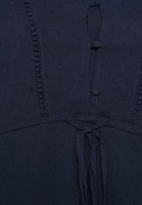 Marineblaue Bluse aus strukturiertem Stoff mit kleinen erhabenen Punkten, Knopfverschluss und einer gebundenen Detail an der Taille. Verzierte Nähte entlang der Säume.