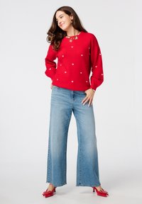 Pull rouge avec des motifs en forme de cœur blancs, assorti à un jean bleu large. Le mannequin porte des chaussures rouges et un collier en or. Textures lisses.