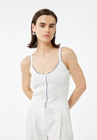 Hvid ribbet tanktop med bølgede blå detaljer og en frontlukning, lavet af et struktureret stof. Modellen poser i lyse bukser.