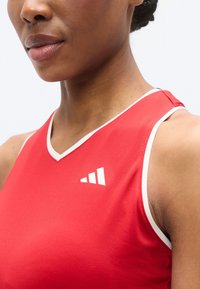 Débardeur de sport rouge avec des accents blancs, présentant un design à col en V et un logo à trois bandes sur la poitrine. Tissu lisse et texturé.