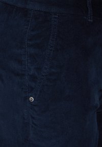 Marineblauwe katoenen broek met een subtiele textuur, voorzien van een zijzak en stikseldetails, benadrukt door een kleine metalen knoop.