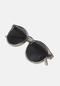 Sonnenbrille mit einem halbtransparenten grauen Rahmen, großen runden schwarzen Gläsern und klaren Bügeln. Minimalistisches Design ohne sichtbare Befestigungen oder Muster.