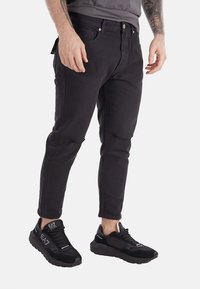 Jeans in denim nero con vestibilità slim, dotati di un design a cinque tasche, cuciture a contrasto e una finitura testurizzata sottile, abbinati a sneakers nere.