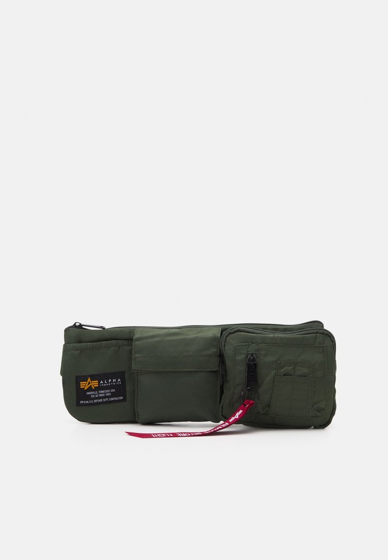Sac banane vert foncé avec trois poches zippées et à rabat, dont une avec un écusson logo Alpha Industries et un curseur de fermeture éclair en tissu rouge.