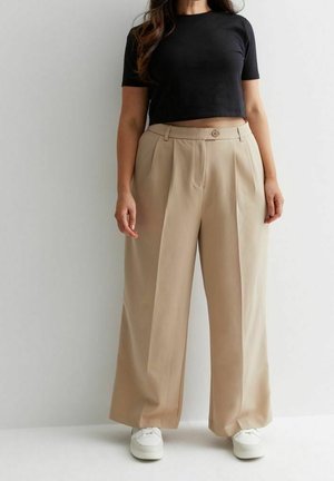 Frau trägt beige weite Hosen, weiße Sneakers und ein schwarzes, kurzärmeliges, bauchfreies Oberteil und steht vor einer schlichten Wand.