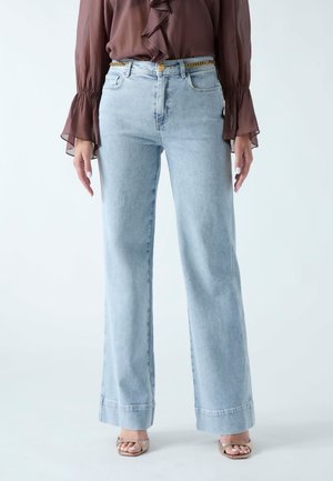 Frau trägt hellblaue Jeans mit weitem Bein, goldene Kettengürtel, braune transparente Langarmbluse und metallisch glänzende offene Sandaletten, stehend vor weißem Hintergrund.