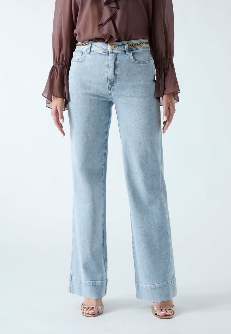 Frau trägt hellblaue Jeans mit weitem Bein, goldene Kettengürtel, braune transparente Langarmbluse und metallisch glänzende offene Sandaletten, stehend vor weißem Hintergrund.