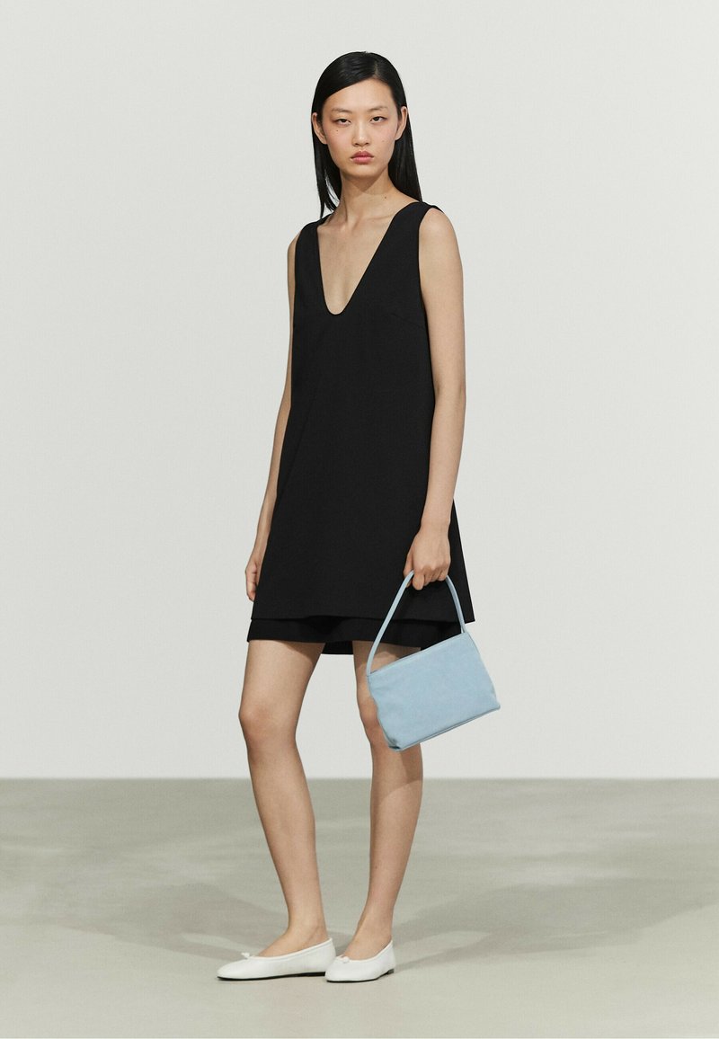 Woman wearing a sleeveless black dress and white flats holding a small light blue handbag standing against a plain light background.