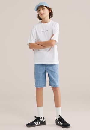 Teen wearing light blue cap, white "Blue Ridge Denim" t-shirt, denim shorts, white socks, and black Adidas sneakers, standing with arms crossed.