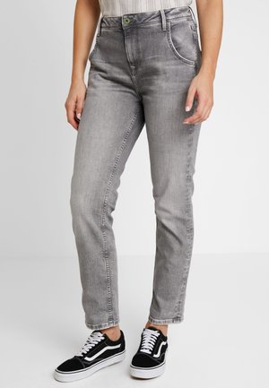 Kvinde iført slidte grå jeans med lige ben og sort-hvide sneakers, stående mod en ensfarvet baggrund.