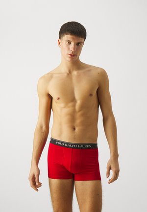 Boxers rojos con una cinturilla de logo negra. Tela suave, diseño ajustado y longitud a medio muslo. El modelo posando erguido con una expresión neutra.