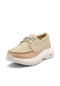Chaussure décontractée beige et tan à lacets avec tige en daim, semelle épaisse blanche texturée et semelle extérieure Vibram pour la traction, vue inclinée vers la gauche sur fond blanc.