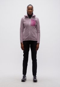 Γυναίκα που στέκεται φορόντας ένα ανοιχτό μωβ μπουφάν με φερμουάρ και τσέπη στο στήθος, μαύρο παντελόνι και παπούτσια για πεζοπορία σε απλό λευκό φόντο.