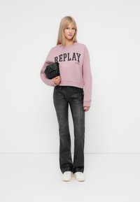 Šviesiai rožinės spalvos trumpa megztinė su "REPLAY" logotipu, derinama su tamsiomis plačiomis džinsinėmis kelnėmis ir baltomis sportbačiais; laiko mažą juodą rankinę.