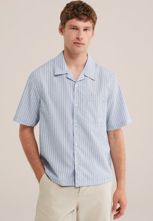 Jeune homme aux cheveux bouclés portant une chemise bleu clair à manches courtes avec des rayures verticales et un pantalon beige clair.