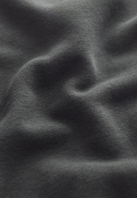 Krupni plan tamnosive tkanine s finom pletenom strukturom i mekim naborima koji stvaraju suptilne sjene.