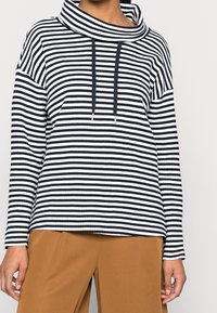 Proužkované tričko s rolákovým výstřihem v námořnické modré a bílé barvě, s volným střihem, dlouhými rukávy a šňůrkami jako detaily. Měkká texturovaná látka.