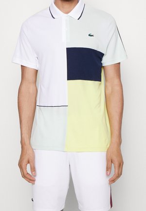 Mann trägt ein kurzärmeliges Polo mit weißen, marineblauen, hellblauen und gelben Blöcken, kombiniert mit weißen Shorts vor schlichtem Hintergrund.